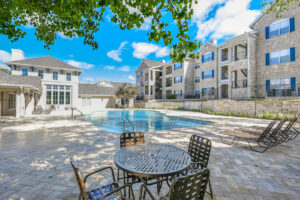 Outdoor pool area at Acclaim at South Congress, featuring lounge chairs, a round patio table, and a view of the apartment buildings and clubhouse.