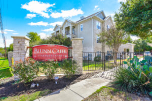 Close-up view of the gated entrance to Acclaim at South Congress with a stone pillar, red signage, and surrounding landscaping.