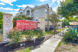 Entrance to Acclaim at South Congress with a red sign reading "Blunn Creek Apartment Homes," surrounded by greenery, flowers, and a gated entrance.