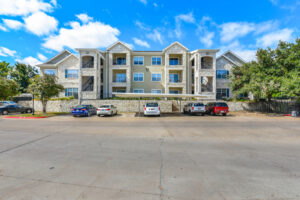 Front view of Acclaim at South Congress apartment complex, showcasing three-story buildings with stone and stucco exteriors, balconies, and covered parking spaces.