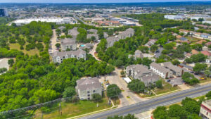 Aerial Exterior wide angle view of Acclaim at South Congress apartment complex, showcasing three-story buildings with stone and stucco exteriors, balconies, and covered parking spaces.