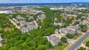 Aerial Exterior wide angle view of Acclaim at South Congress apartment complex, showcasing three-story buildings with stone and stucco exteriors, balconies, and covered parking spaces.