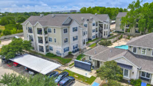 Aerial Exterior wide angle view of Acclaim at South Congress apartment complex, showcasing three-story buildings with stone and stucco exteriors, balconies, and covered parking spaces.