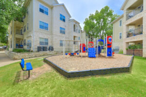 Children's playground area within Acclaim at South Congress, featuring slides, climbing structures, and benches, surrounded by apartment buildings.
