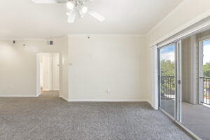 Spacious living area with carpeted flooring, white walls, ceiling fan, and a sliding glass door leading to a balcony.