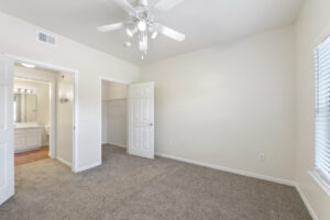 Spacious living area with carpeted flooring, white walls, ceiling fan, and a door leading to bathroom.
